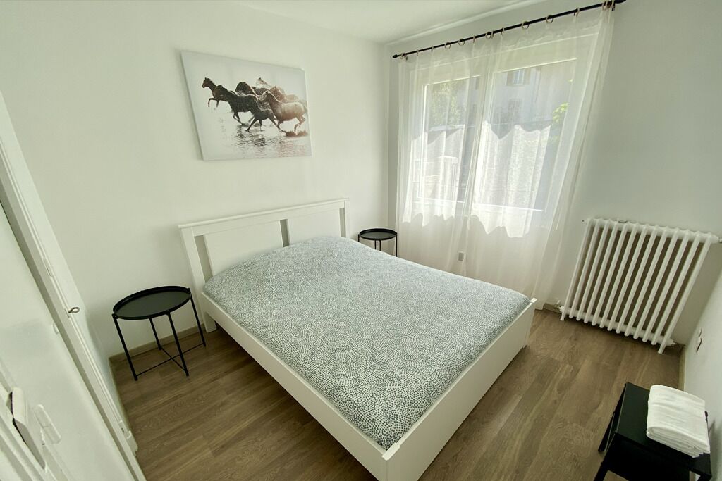 A bedroom with a bed, a small table, a radiator, and a window with curtains.
