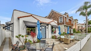 Ein Patio mit einem runden Tisch und Stühlen, einem weißen Zaun sowie einem Haus mit blauer Tür und braunen Dachziegeln.