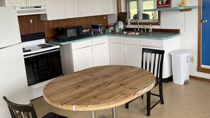 Una cucina funzionale con mobili bianchi, un fornello, un forno a microonde e un lavello. Si nota un tavolo da pranzo in legno con sedie, un cestino per i rifiuti e una sveglia appesa al muro.
