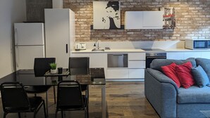 Une cuisine moderne avec des meubles blancs, un mur en briques et un coin repas comprenant une table en verre et des chaises noires.