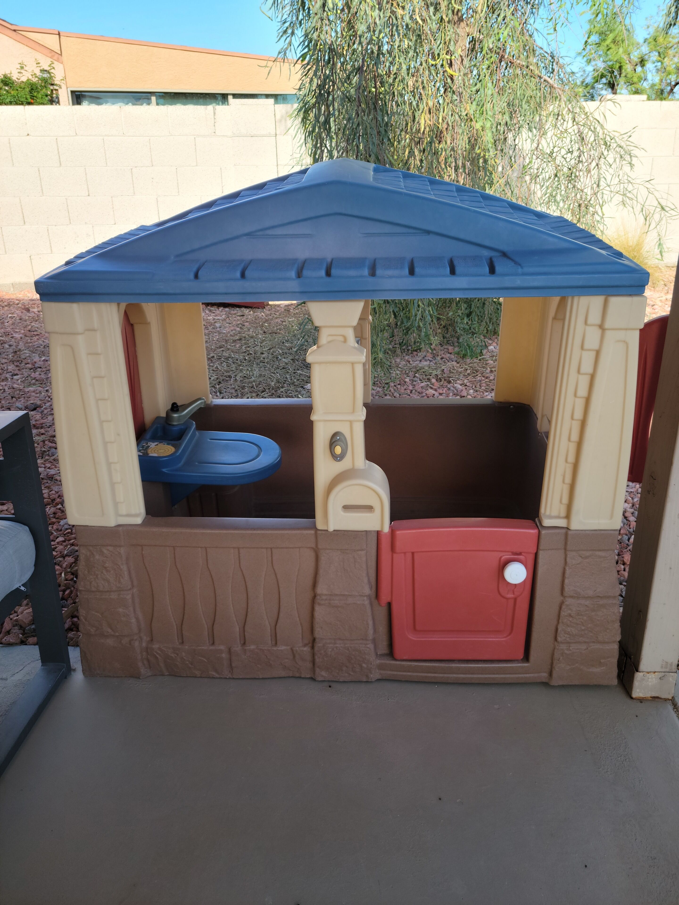 A children's playhouse with a blue roof, beige walls, and a red door.