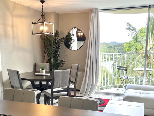 A modern dining area with a round table, grey upholstered chairs, a chandelier, and a large mirror. A balcony with a white railing and outdoor furniture is visible.