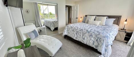 A bedroom with a bed, a chair, a desk, a TV, and a balcony view.