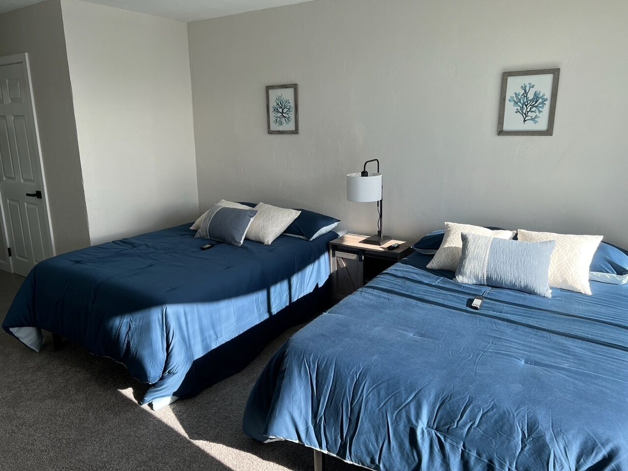 A room with two beds, each with blue bedding and white pillows. There are two framed pictures on the wall, a bedside table with a lamp, and a door on the left.