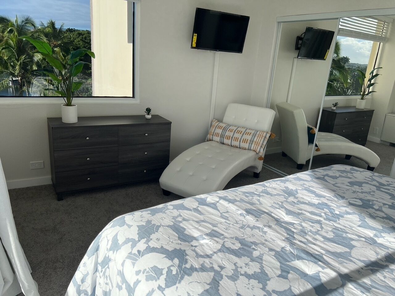 A bedroom with a bed, a dresser, a chair, a TV, and a window with a view of palm trees.