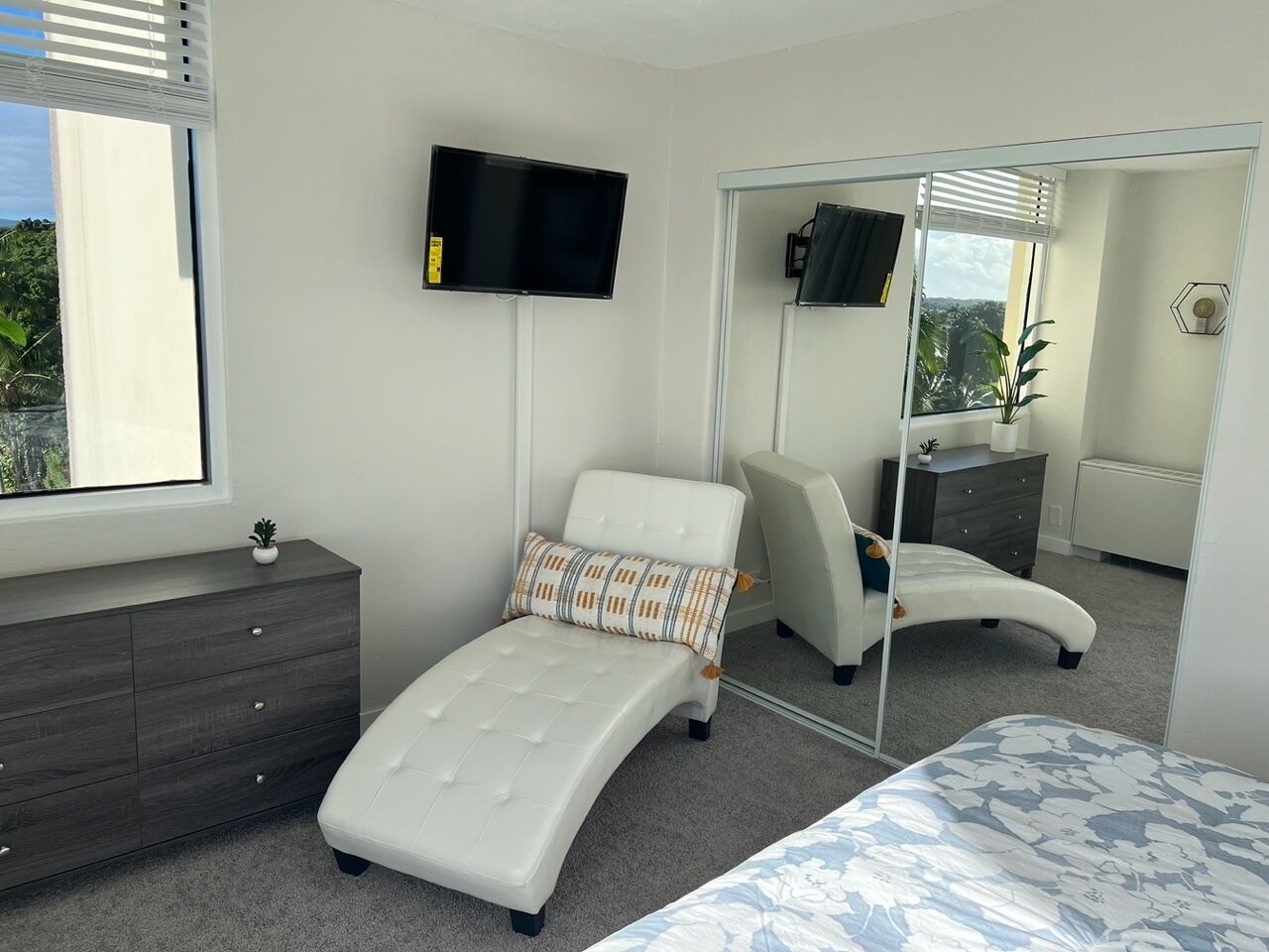 Een moderne slaapkamer met een bed, een kaptafel met spiegel, een televisie aan de muur en een raam met jaloezieën.