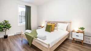 Une chambre à coucher bien rangée, avec un lit en bois, une table de chevet avec une lampe, une petite plante en pot et une fenêtre avec des rideaux.