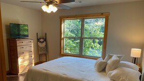 Ein Schlafzimmer mit einem großen Bett, einem Schminktisch, einem Fernseher, einem Fenster mit Blick auf Bäume und einer Lampe.