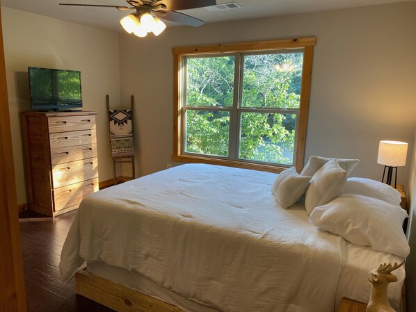 A bedroom with a large bed, a dresser, a TV, a window with a view of trees, and a lamp. - Blue Ridge Cabin within 40 feet of the Toccoa River and Trout Fishing (Blue Ridge)