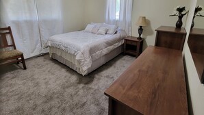 A bedroom with a bed, a wooden nightstand, a wooden desk, a chair, and a lamp.