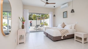 A bedroom with a bed, bedside tables, a ceiling fan, an air conditioner, a large mirror, and a sliding glass door leading to a balcony with outdoor furniture. - Villa Varia (Rarotonga)