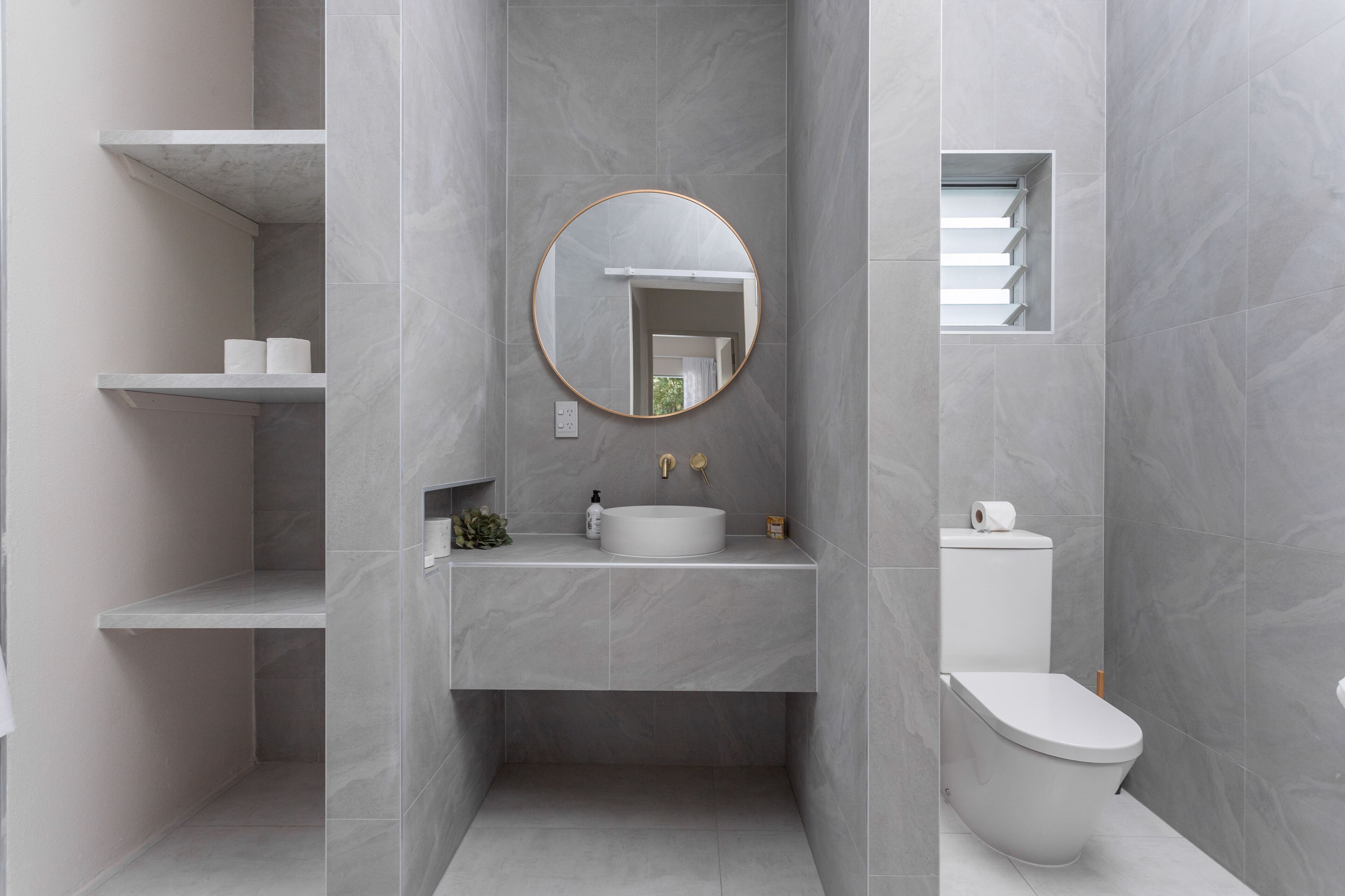 A modern bathroom with a round mirror, white sink, and toilet. Shelves are mounted on the wall, and a small plant is placed on the sink counter.
