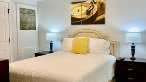 Un dormitorio con una cama grande, dos mesitas de noche con lámparas, un ventilador de techo y una obra de arte colgada en la pared.