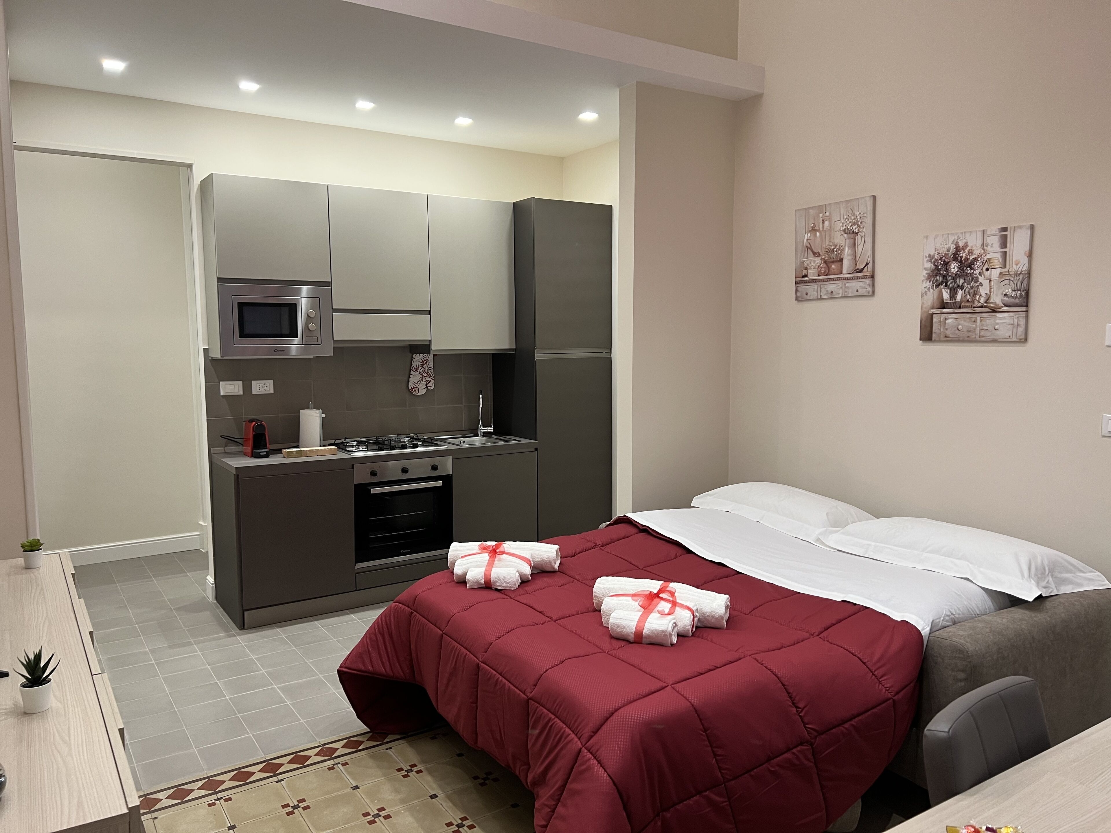 Habitación de hotel pequeña con kitchenette, una cama con ropa de cama roja y dos obras de arte colgadas en la pared.