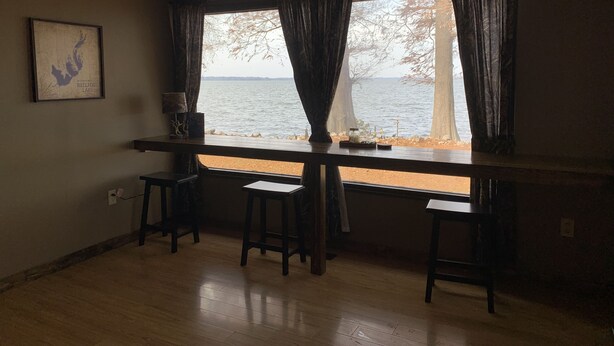 Una habitación con un ventanal grande que da a un lago, suelo de madera y dos taburetes de bar.