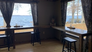 Una habitación con una mesa de madera y dos sillas, una ventana con vista a un lago y árboles, y una estantería con frascos y una figurita.