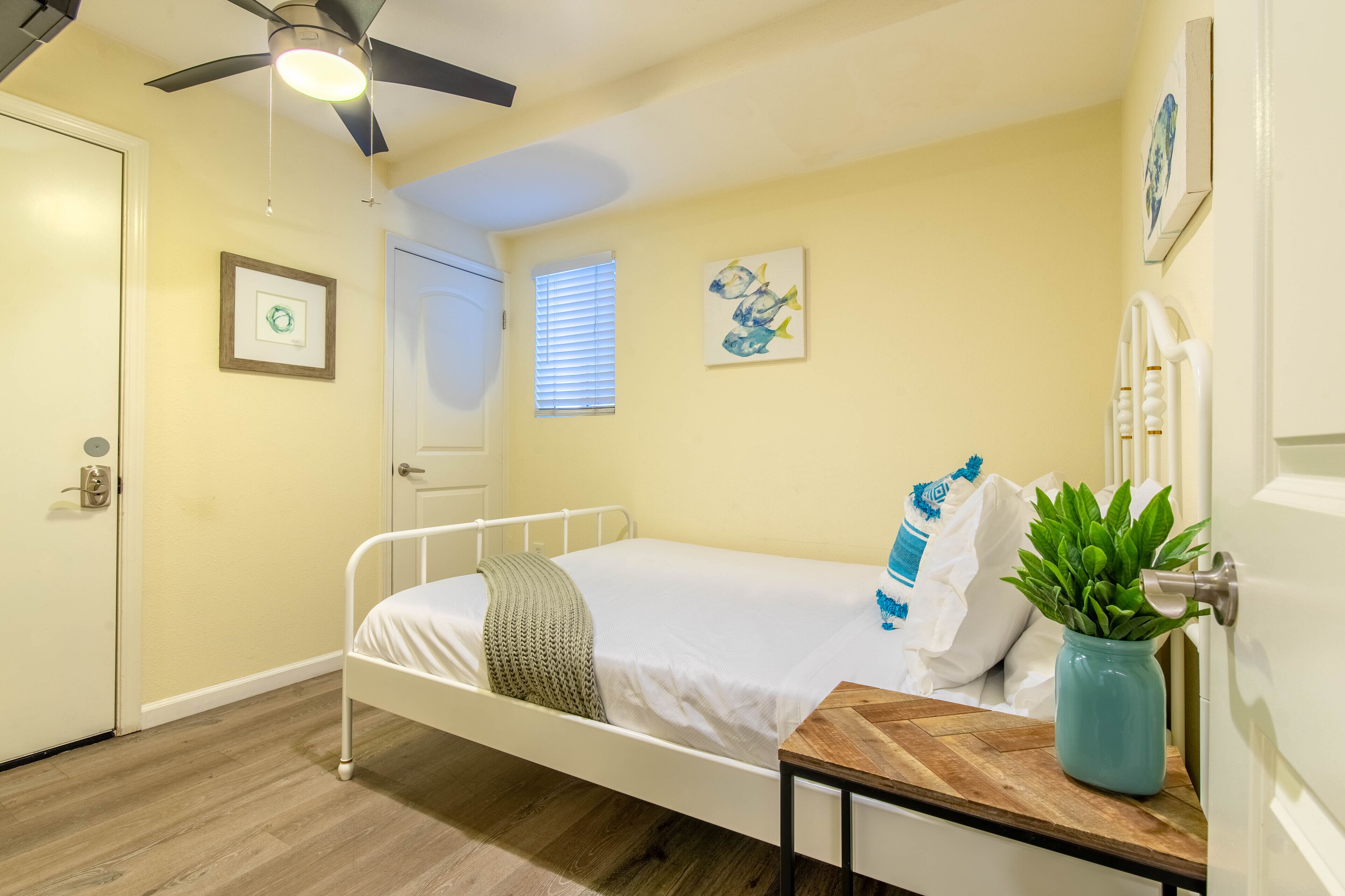 Une chambre à coucher comprenant un lit, un ventilateur de plafond, une fenêtre avec des stores, une porte et un mur orné de tableaux.