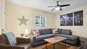 A living room with a grey sectional sofa, a wooden coffee table, a ceiling fan, a starfish decoration, a window with blinds, and three cityscape paintings. - Ocean View Perfection - Parking, W/D, Pet Friendly & Steps to Sand (Mission Beach)