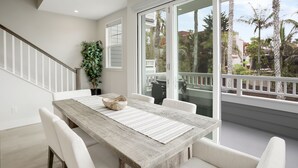 A modern dining area with a wooden table, white chairs, and a large window with a view of palm trees and buildings. - Designer Luxury Coastal Home with Panoramic Ocean Views, Rooftop Deck, & Fire Pit (La Jolla)