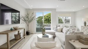 A modern living room with a large flat-screen TV, a sofa set, a coffee table, and a potted plant. - Designer Luxury Coastal Home with Panoramic Ocean Views, Rooftop Deck, & Fire Pit (La Jolla)