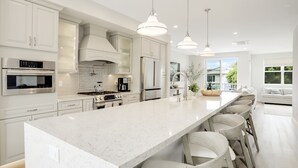 A modern kitchen with white cabinetry, stainless steel appliances, and a large island with bar stools. - Designer Luxury Coastal Home with Panoramic Ocean Views, Rooftop Deck, & Fire Pit (La Jolla)