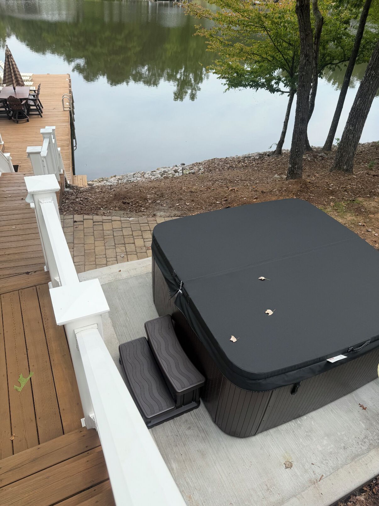 Een hottub op een houten terras met een witte reling, met uitzicht op een waterpartij en bomen.
