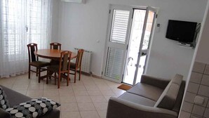 A living room with a sofa, a dining table with chairs, a TV mounted on the wall, and a sliding glass door leading to an outdoor area.