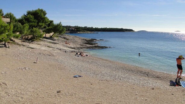 A pebbled beach with clear blue water, a few people sunbathing, and a distant shoreline with buildings.