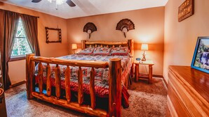 Een slaapkamer met een houten bed, een plafondventilator, twee wandlampen, een lijst met een afbeelding en een raam met gordijnen.