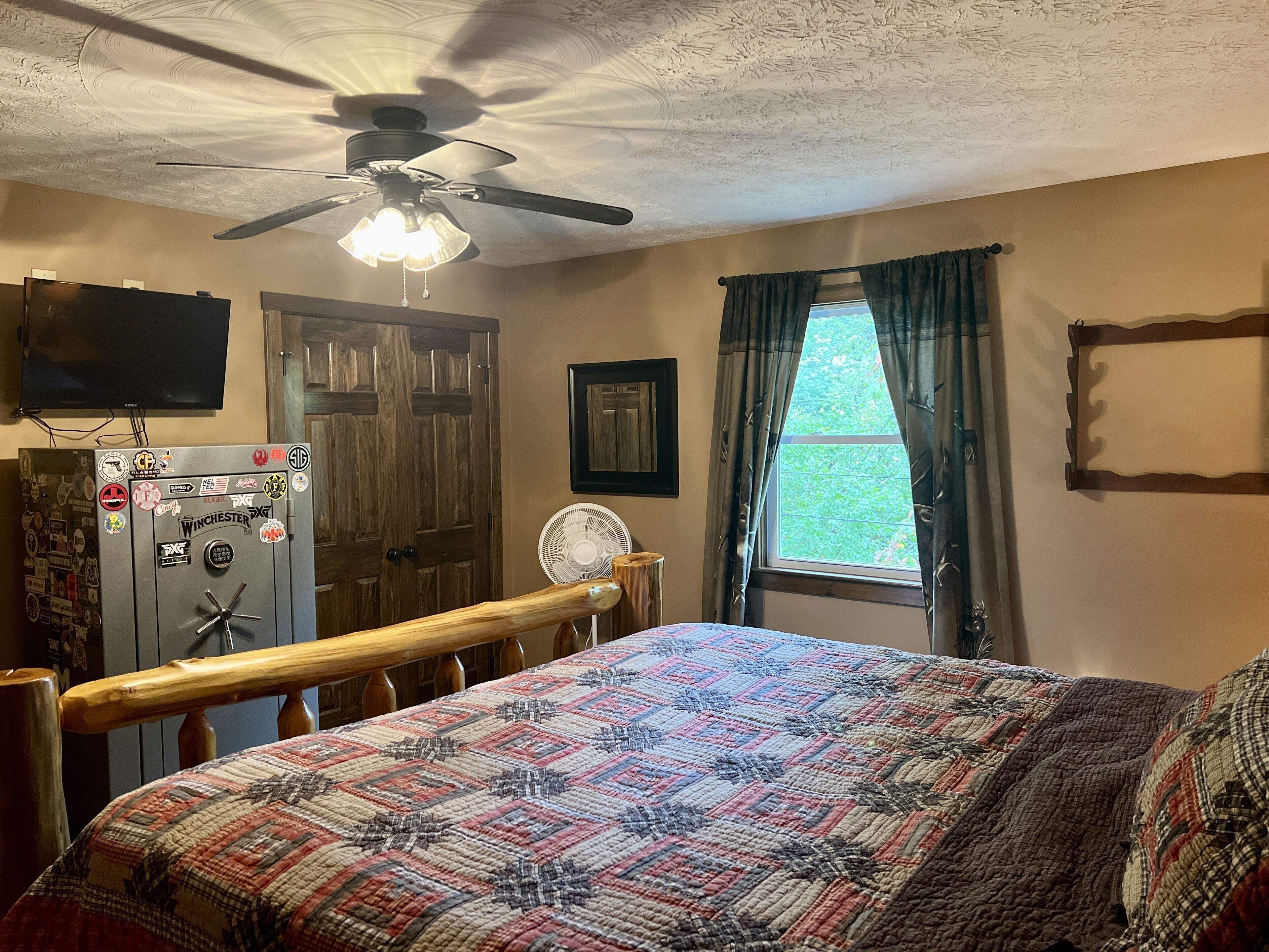 Ein Schlafzimmer mit einem Bett, einem Deckenventilator, einem Fenster mit Vorhängen, einem an der Wand montierten Fernseher und einem Safe mit verschiedenen Aufklebern.