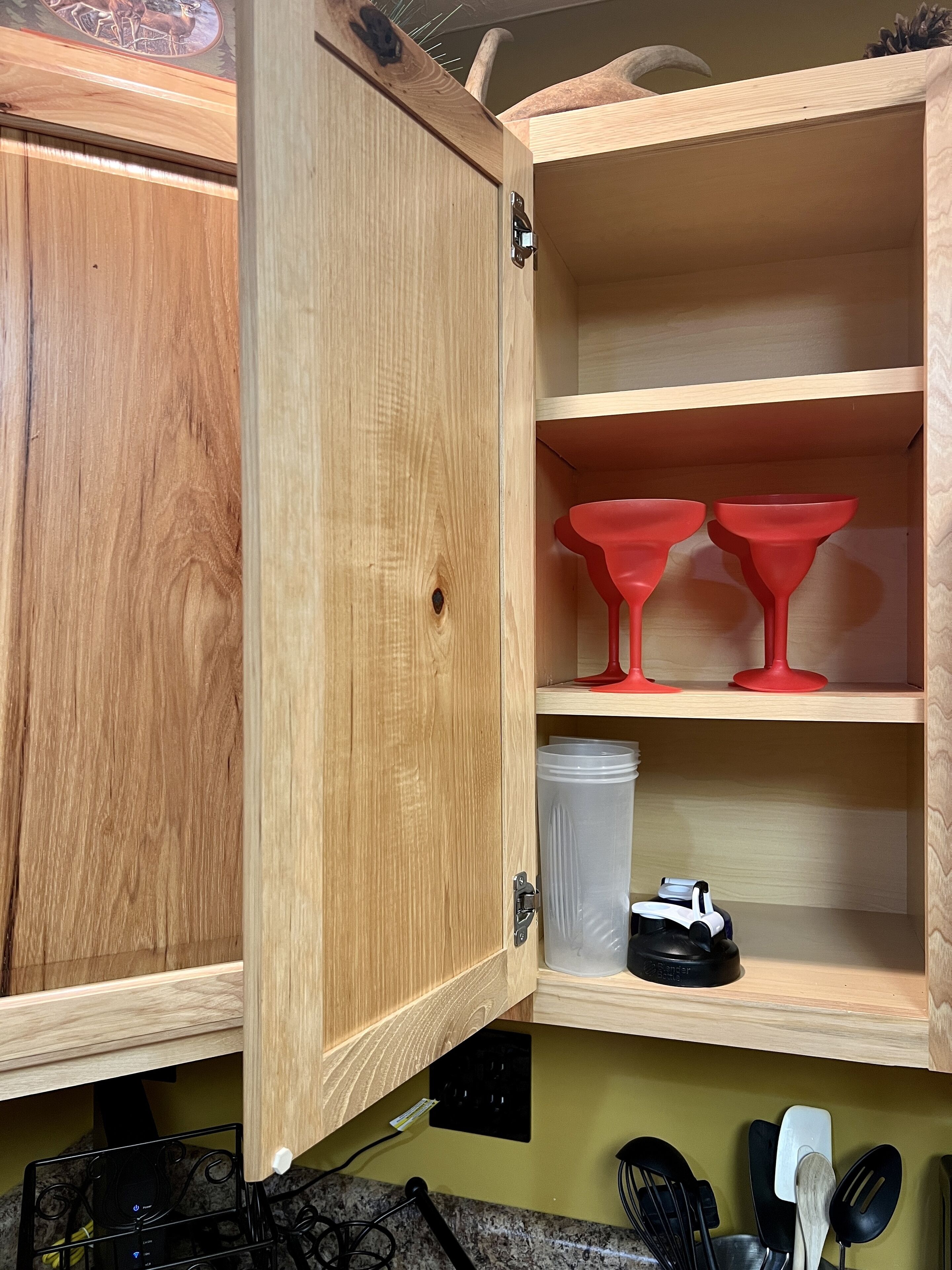 Un meuble en bois avec une porte à charnières, une étagère contenant des ustensiles de cuisine rouges, un mixeur sur l’étagère inférieure et un plan de travail de cuisine avec divers ustensiles.