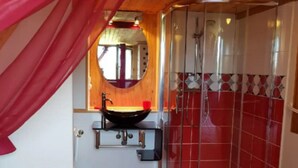A bedroom with a bed, a red curtain, a shower with a glass door, a sink, and a mirror.