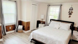 A bedroom with a large bed, wooden floor, white walls, a nightstand, a lamp, and a dreamcatcher on the wall. - Beautiful Cottage! 2.5m from I-95,13m to FtBragg (Fayetteville)