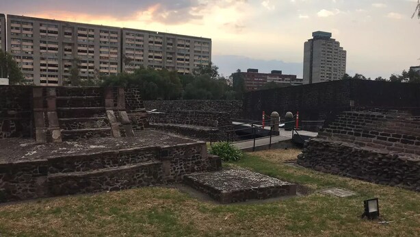 Un site archéologique comprenant des structures et des escaliers en pierre, entouré de bâtiments modernes.