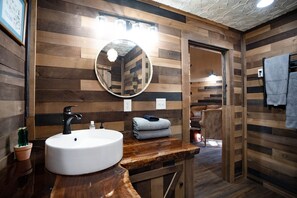 Une salle de bain aux murs lambrissés, avec un miroir rond, un lavabo blanc et un plan de toilette en bois.