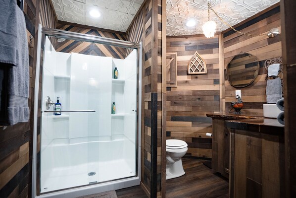 Baño con ducha de vidrio, paneles de madera, inodoro blanco y tocador de madera con espejo.