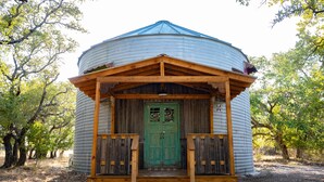 Un bâtiment rustique de forme cylindrique, doté d’un porche en bois et d’une porte verte, entouré d’arbres.