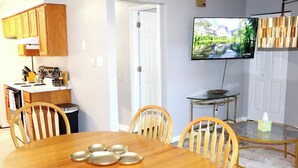 Un comedor con una mesa y sillas de madera, una cocina con armarios de madera y estufa, un televisor montado en la pared y una pequeña mesa de vidrio con una caja de pañuelos.