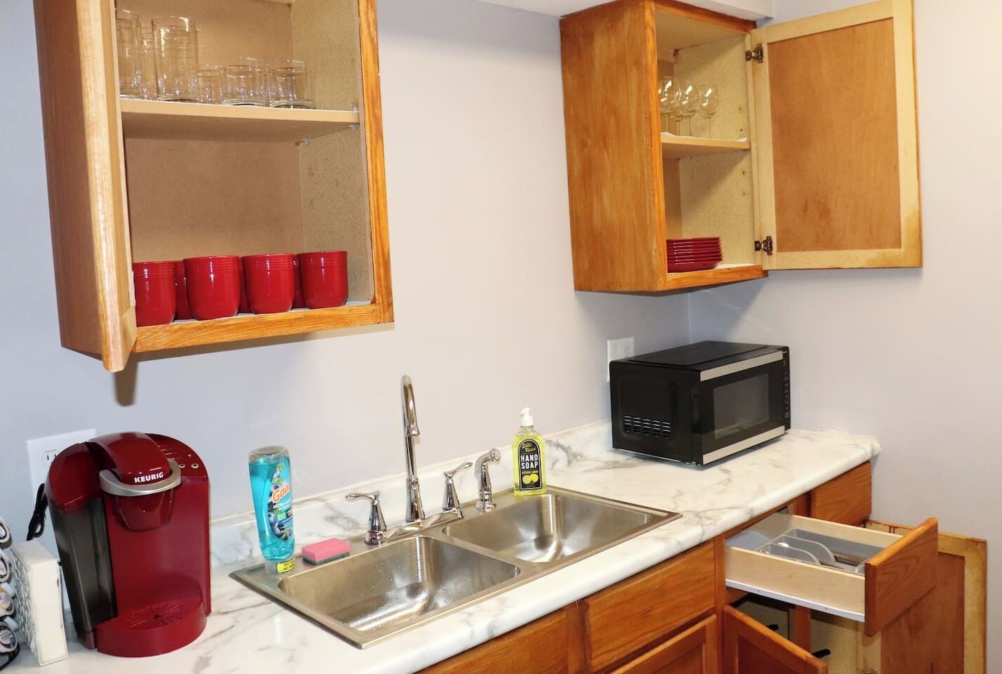 A compact kitchen with a sink, a coffee maker, a microwave, and cabinets with glassware and red cups.
