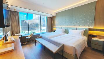 A modern hotel room with two beds, a desk, a chair, and a view of the cityscape.