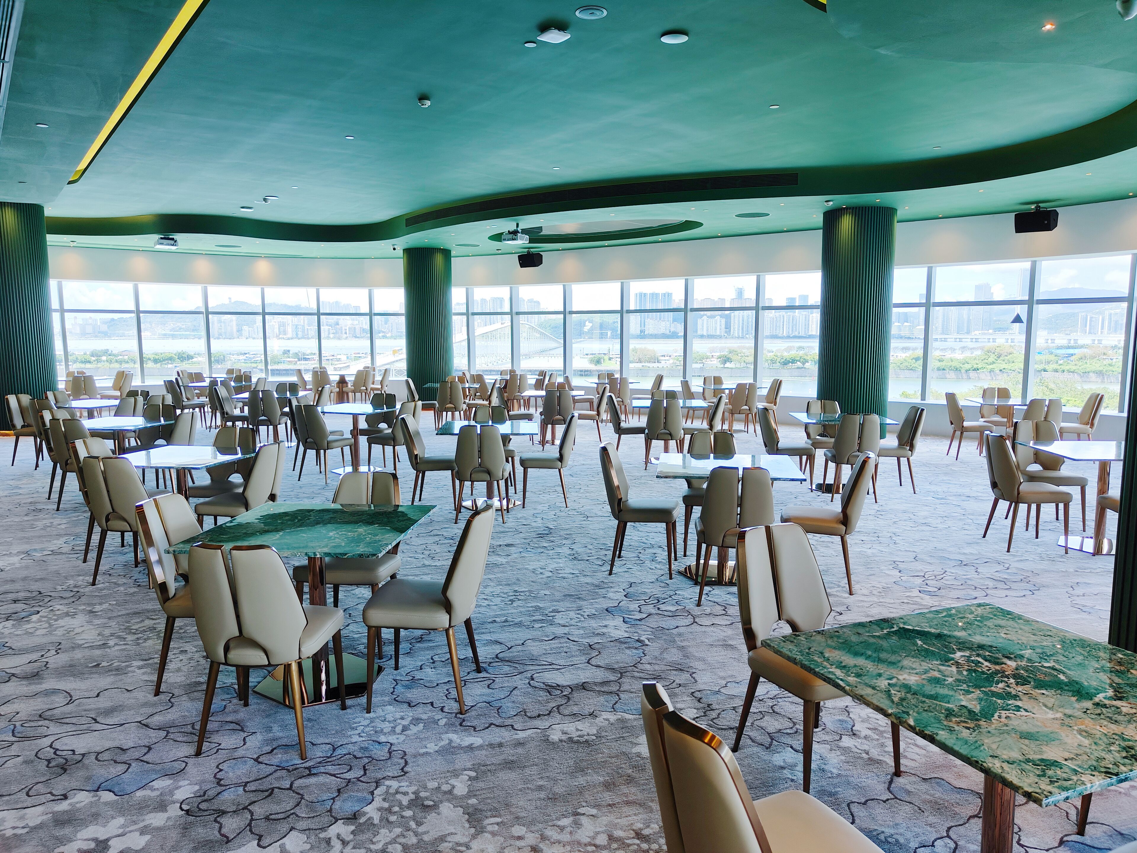 A spacious indoor dining area with tables and chairs arranged for groups, large windows offering a view of the cityscape, and a modern ceiling design.