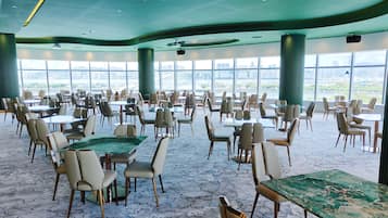 A spacious indoor dining area with tables and chairs arranged for groups, large windows offering a view of the cityscape, and a modern ceiling design.