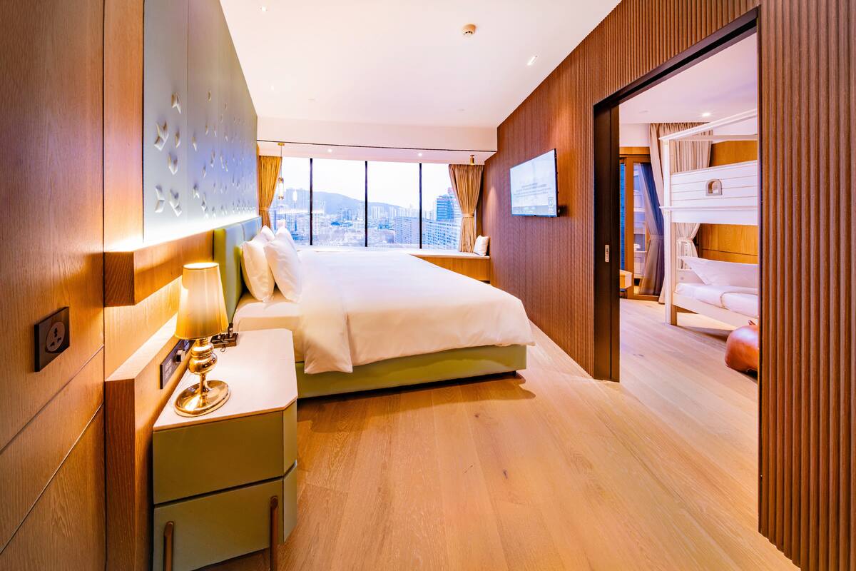 Une chambre d’hôtel moderne dotée d’un grand lit, d’une table de chevet avec une lampe, d’un téléviseur fixé au mur et offrant une vue sur la ville grâce à de grandes fenêtres.