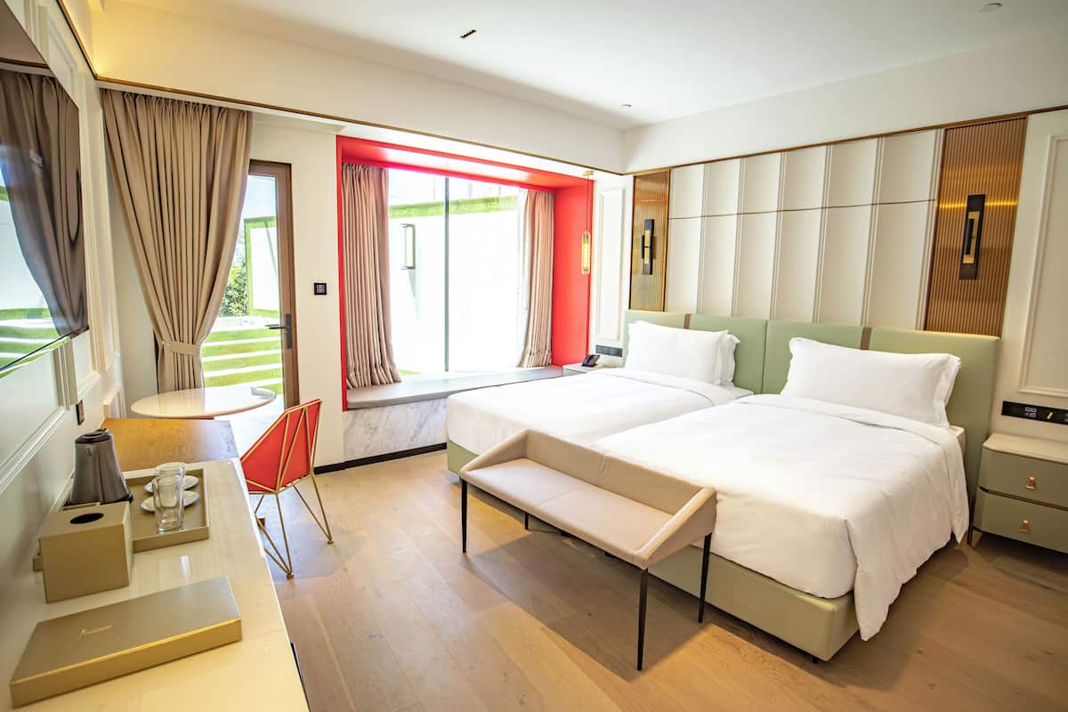 Une chambre d’hôtel moderne avec deux lits, une petite table avec une lampe, un banc et une vue sur un jardin par la fenêtre.