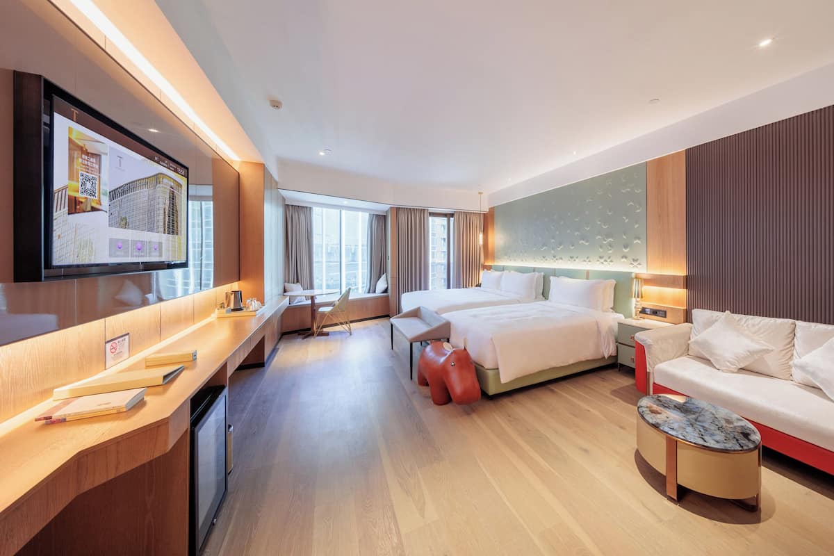 Une chambre d’hôtel moderne, dotée d’un grand lit, d’un canapé, d’un bureau et d’un téléviseur diffusant des images d’un paysage urbain.