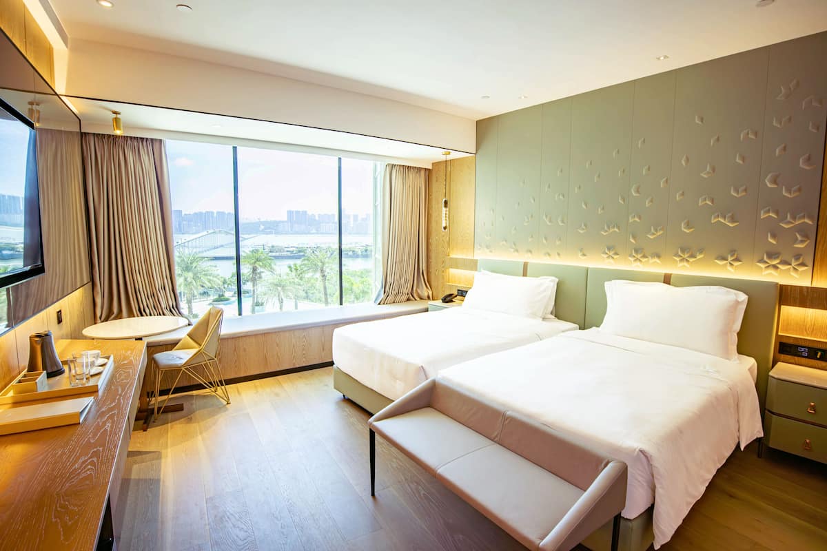 Une chambre d’hôtel avec deux lits, un bureau avec une chaise, une grande fenêtre donnant sur un paysage urbain et un mur orné d’un motif texturé.