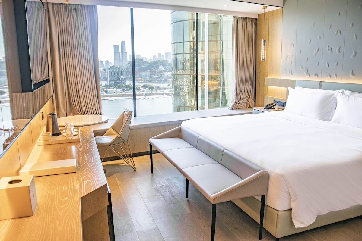 Une chambre d’hôtel moderne dotée d’un grand lit, d’un bureau avec une chaise, d’un banc et offrant une vue sur le paysage urbain grâce à de grandes fenêtres.