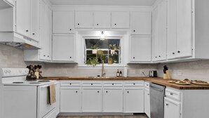 A modern kitchen with white cabinets, a wooden countertop, and a window with plants.