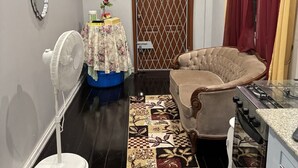A compact living space with a sofa, a dining table, a kitchen area, a standing fan, and a decorative rug. - In City - Minutes to Regent St, Botanical Gardens, Cuffy Sq, and Bourda Market.  (Georgetown)