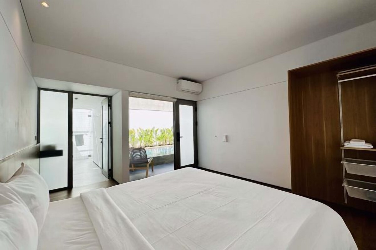 Une chambre d’hôtel moderne avec un grand lit, une armoire en bois et une porte-fenêtre coulissante donnant sur un balcon avec vue sur la verdure.